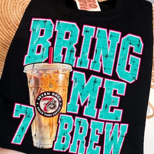 May include: A black crewneck sweatshirt featuring a graphic of an iced coffee in a clear plastic cup with a red straw. The text BRING ME 7 BREW is printed in large, distressed teal block letters with pink outlines. A Seven Brew logo is on the cup.