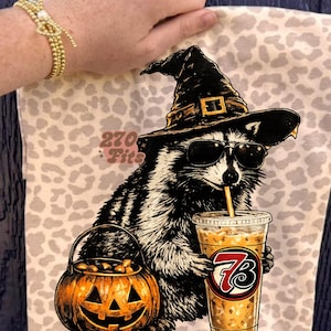 May include: A beige tote bag with a leopard print pattern featuring a graphic of a raccoon wearing a witch hat and sunglasses, drinking from a cup with a straw. The raccoon holds a jack-o'-lantern bucket. The text 270 Fits is printed in pink.