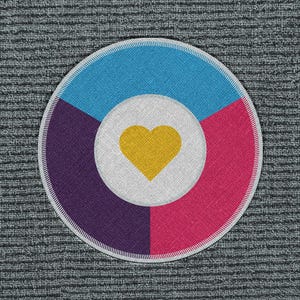 May include: Circular patch with a segmented design. The top section is light blue, the right is pink, and the left is purple. A white circle in the center contains a yellow heart. The patch has a white border and is set against a textured gray background.