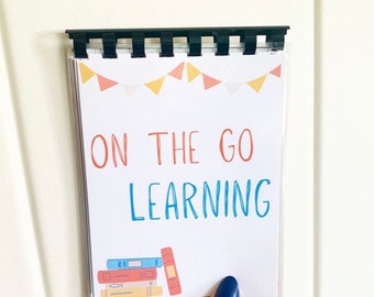 Pode incluir: Um pequeno caderno branco com uma encadernação em espiral preta. A capa apresenta uma ilustração colorida de uma pilha de livros e o texto "ON THE GO LEARNING".