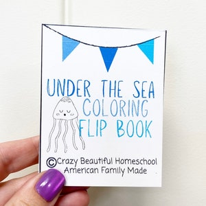 May include: A white card with blue and white bunting at the top. The card says "Under the Sea Coloring Flip Book" with a black and white illustration of a jellyfish. The bottom of the card says "&copy;Crazy Beautiful Homeschool American Family Made."