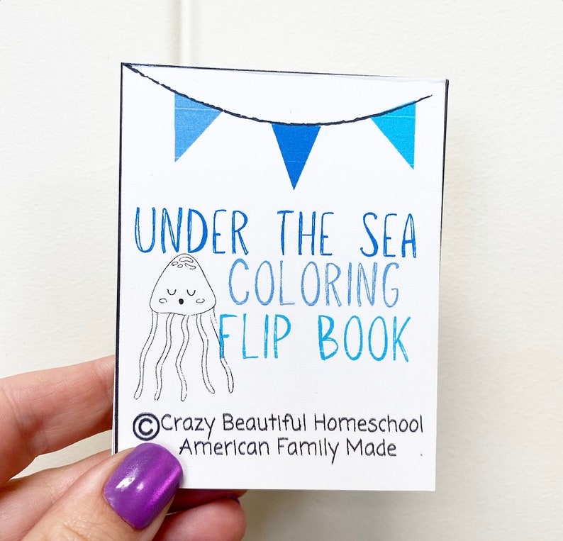 May include: A white card with blue and white bunting at the top. The card says "Under the Sea Coloring Flip Book" with a black and white illustration of a jellyfish. The bottom of the card says "&copy;Crazy Beautiful Homeschool American Family Made."