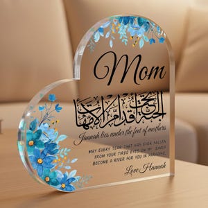 May include: A heart-shaped acrylic plaque with floral accents and the word "Mom" in elegant script. The plaque features Arabic calligraphy and English text that reads "Jannah lies under the feet of mothers." A personalized message, "Love Hannah," is also included.