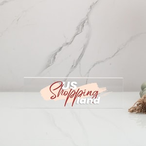 May include: A clear acrylic sign with the text "US Shopping Land" in white and red lettering. The text is on a pink brushstroke background.