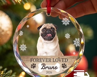 May include: A round, faceted glass Christmas ornament featuring a portrait of a tan pug dog. The ornament includes white snowflake graphics, a wood-textured banner with the text "FOREVER LOVED", a bone shape with the name "Bruno", and the year "2026".