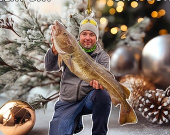 May include: A Christmas ornament featuring a man holding a large fish. The ornament is made of clear acrylic and has a gold string for hanging. The man is wearing a gray jacket, a black hat, and blue pants. The fish is white and has a dark stripe down its side.