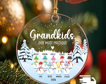 May include: A round, clear acrylic Christmas ornament featuring the text "Grandkids OUR MOST PRECIOUS" above a snowy landscape with pine trees and ten illustrated snowmen wearing colorful hats and scarves, each labeled with a name, plus the year 2026.