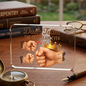 May include: A clear acrylic plaque with the words "Dad's Team THE BEST TEAM EVER" in gold lettering. The plaque features illustrations of fists, a gold crown, and the names "Liam" and "Noah".