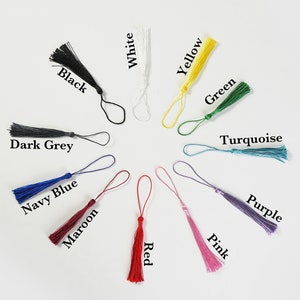 May include: Assortment of 12 tassel keychains in various colors, including black, white, yellow, green, turquoise, purple, pink, navy blue, maroon, red, and dark grey. Each tassel is attached to a small loop.