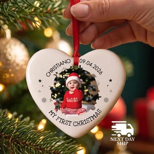May include: A white heart-shaped ceramic ornament hanging from a red ribbon. The ornament features a photo of a baby in a red outfit, the name Christiano, the date 09.04.2026, and the text My First Christmas, accented with small black stars and snowflakes.