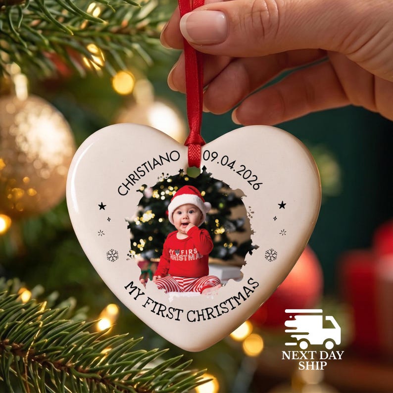 May include: A white heart-shaped ceramic ornament hanging from a red ribbon. The ornament features a photo of a baby in a red outfit, the name Christiano, the date 09.04.2026, and the text My First Christmas, accented with small black stars and snowflakes.