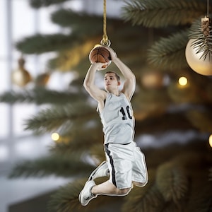 May include: A basketball-themed Christmas ornament. The ornament depicts a basketball player in a gray jersey with the number 10, shooting a basketball. The ornament is hanging from a gold string against a Christmas tree background.