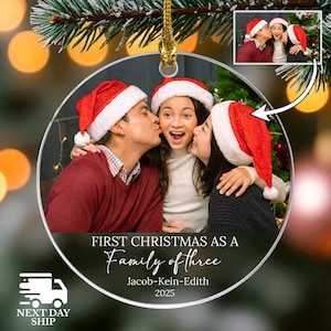 May include: A clear, round Christmas ornament with a family photo inside. The family of three is wearing Santa hats and kissing. Text on the ornament reads "First Christmas as a Family of Three" with the family's names and the year 2025.