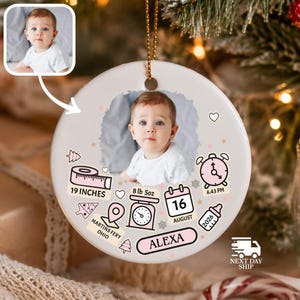 K&ouml;nnte beinhalten: Ein rundes, wei&szlig;es Keramikornament mit einem zentralen Babyfoto. Die Grafiken zeigen ein Ma&szlig;band mit der Aufschrift 19 INCHES, eine Waage mit 8 lb 5oz, einen Kalender mit 16 AUGUST, eine Uhr auf 6:43 PM, ein Babyfl&auml;schchen und den Namen ALEXA.