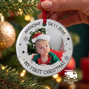 May include: A round white ceramic Christmas ornament featuring a central photograph of a baby wearing a Santa hat and green clothing. The ornament is printed with the text ANTHONY, 04.11.2026, and MY FIRST CHRISTMAS, accented by black snowflakes and stars.