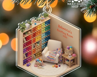May include: A clear acrylic ornament shaped like a room with a bookshelf filled with colorful yarn, a white armchair, and a small table. The text "Rebecca's Knotting Room 2025" is visible. The ornament is hanging from a gold string.