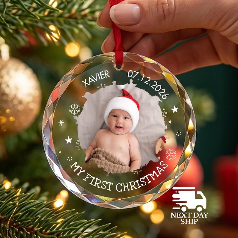 May include: A circular, faceted glass Christmas ornament featuring a central photograph of a baby wearing a red and white Santa hat. The ornament is printed with the name XAVIER, the date 07.12.2026, and the text MY FIRST CHRISTMAS in white.