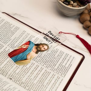 May include: A clear plastic bookmark with a picture of Jesus Christ and the text "David! Is That SMUT?" printed on it. The bookmark has a red tassel.