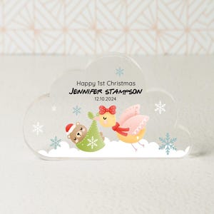 May include: A white cloud-shaped acrylic ornament with a Christmas-themed design. It features a cartoon bird carrying a baby bear in a green and red stocking. The text "Happy 1st Christmas Jennifer Stampson 12.10.2024" is printed on the ornament.