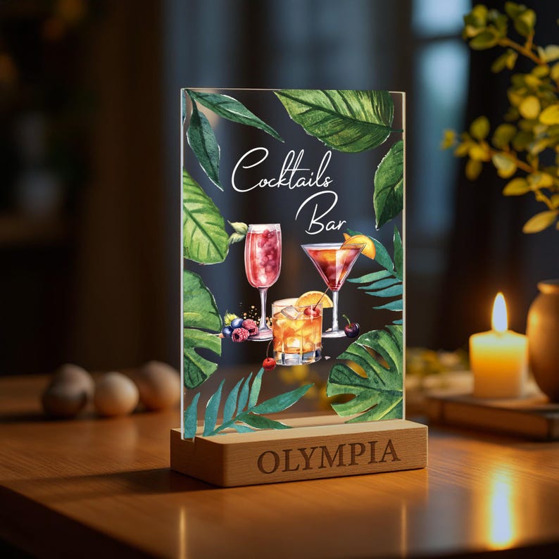 May include: A clear acrylic sign with a tropical leaf design and three cocktails. The sign reads "Cocktails Bar" and has a wooden base with the word "OLYMPIA" engraved on it.