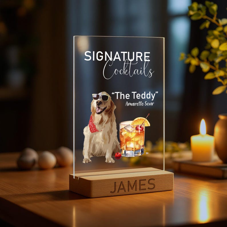 May include: A clear acrylic sign with the text "SIGNATURE Cocktails" and "The Teddy" Amaretto Sour. The sign features a cartoon illustration of a golden retriever wearing sunglasses and a red bandana. The sign is on a wooden base with the name "JAMES" engraved on it.