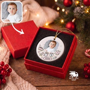 K&ouml;nnte beinhalten: Ein rundes, wei&szlig;es Keramikornament in einer roten Geschenkbox mit einem Babyfoto, dem Namen ALEXA und Geburtsdaten wie 19 INCHES, 16 AUGUST und 2026, verziert mit kleinen Strichzeichnungen einer Waage, einer Uhr und einer Babyflasche.