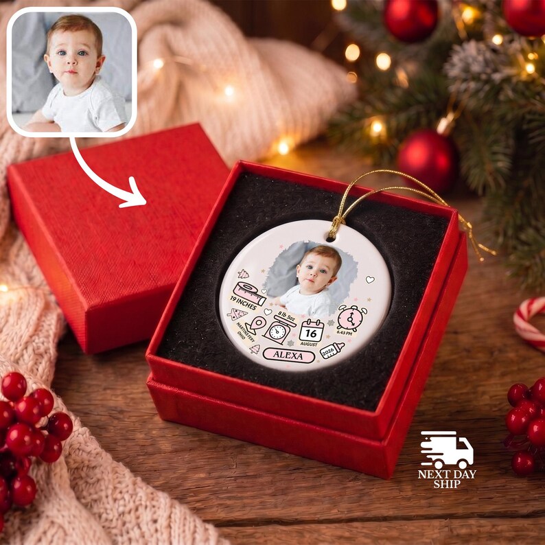 K&ouml;nnte beinhalten: Ein rundes, wei&szlig;es Keramikornament in einer roten Geschenkbox mit einem Babyfoto, dem Namen ALEXA und Geburtsdaten wie 19 INCHES, 16 AUGUST und 2026, verziert mit kleinen Strichzeichnungen einer Waage, einer Uhr und einer Babyflasche.