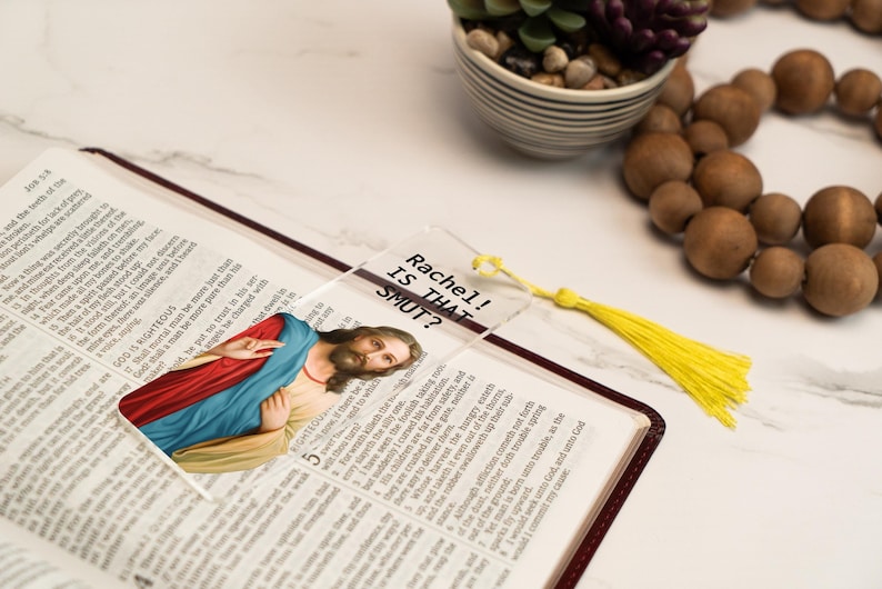 May include: A clear acrylic bookmark with a picture of Jesus Christ and the text "Rachel! Is That Smut?" printed on it. The bookmark has a yellow tassel.