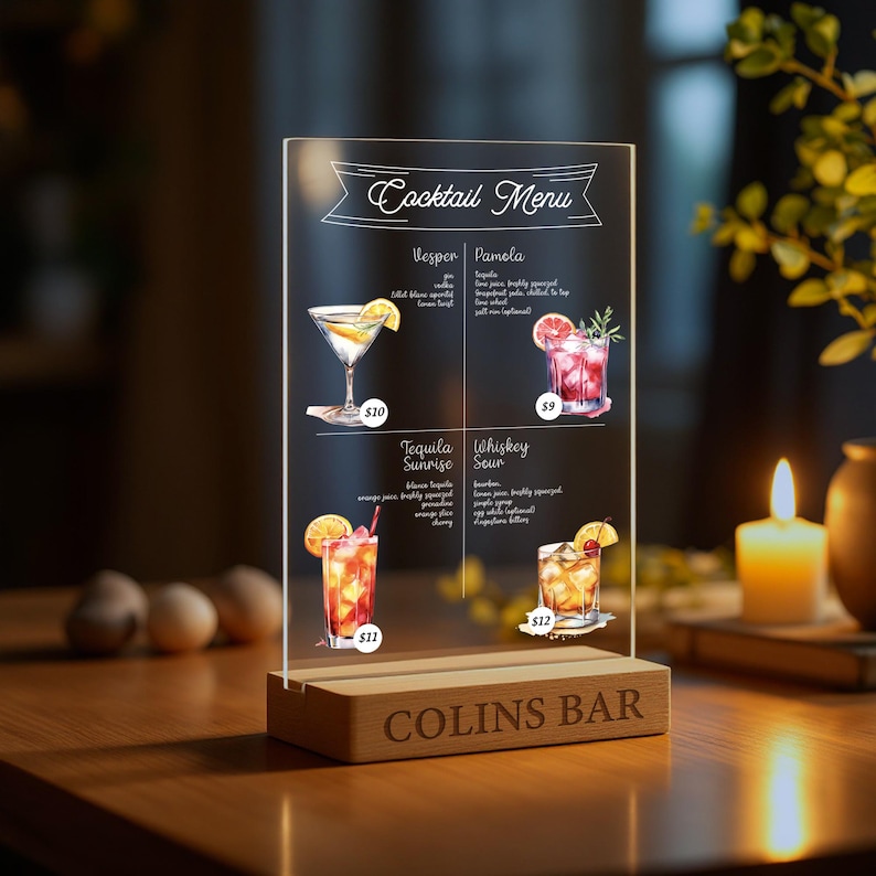 May include: A clear acrylic sign with a wooden base that reads "COLINS BAR" and displays a cocktail menu with four different drinks: Vesper, Pamola, Tequila Sunrise, and Whiskey Sour. Each drink is illustrated with a watercolour painting and priced.