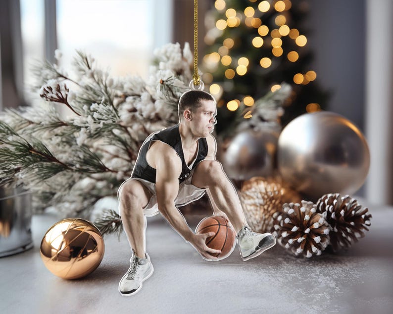 May include: A basketball-themed Christmas ornament featuring a male basketball player in a black and white uniform dribbling a basketball. The ornament is hanging with a gold string, with a blurred Christmas tree and ornaments in the background.