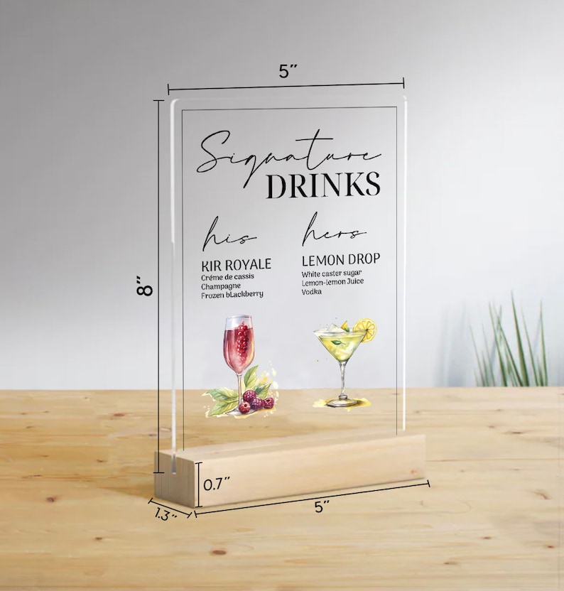 May include: A clear acrylic sign with a wooden base, measuring 8 inches tall and 5 inches wide. The sign features the text "Signature Drinks" with the words "his" and "hers" above two drink recipes. The "his" drink is called "Kir Royale" and the "hers" drink is called "Lemon Drop".