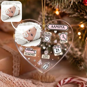 K&ouml;nnte beinhalten: Ein herzf&ouml;rmiges, transparentes Acryl-Ornament mit einem Foto eines Babys mit wei&szlig;em Stirnband und Schnuller. Das Ornament zeigt den Namen AAMINA, eine Waage mit 8 lb 5oz, eine Uhrzeit von 13:59 Uhr, ein Ma&szlig;band mit 19 INCHES und 21 JUNI.