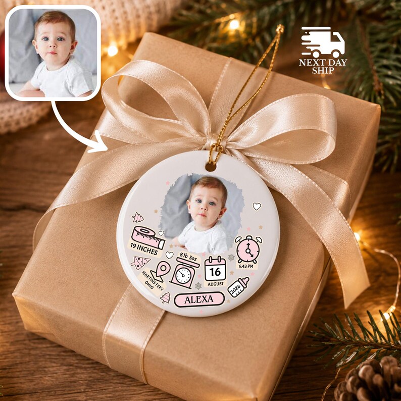 K&ouml;nnte beinhalten: Ein rundes, wei&szlig;es Keramikornament mit einem zentralen Babyfoto, umgeben von gedruckten Symbolen wie einem Ma&szlig;band, einer Waage, einer Uhr, einem Kalender mit dem Datum 16 AUGUST und dem Namen ALEXA, verziert mit einem hellgoldenen Band.