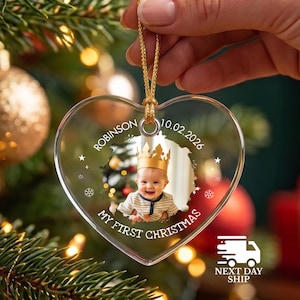 May include: A heart-shaped clear acrylic ornament featuring a central photograph of a baby wearing a gold crown. The text reads "ROBINSON", "10.02.2026", and "MY FIRST CHRISTMAS" with small star and snowflake graphics. It hangs from a gold cord.