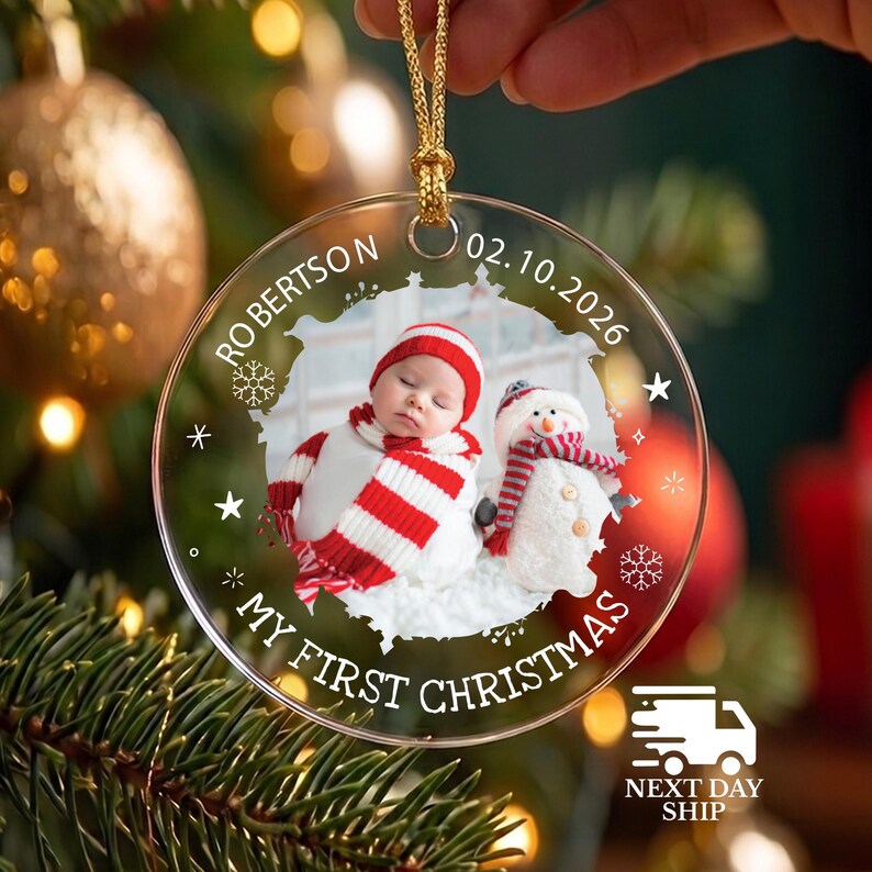 May include: A clear circular acrylic Christmas ornament featuring a photo of a baby in a red and white striped hat and scarf next to a snowman. The text reads "ROBERTSON", "02.10.2026", and "MY FIRST CHRISTMAS" with white snowflake and star accents.