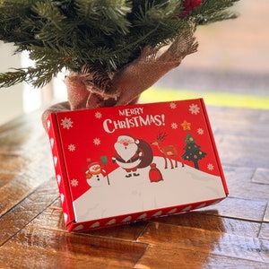 K&ouml;nnte beinhalten: Eine rechteckige rote Geschenkbox aus Karton mit dem Schriftzug Merry Christmas in wei&szlig;en Buchstaben. Die Box zeigt eine festliche grafische Illustration mit dem Weihnachtsmann, einem Schneemann, einem Rentier und einem Weihnachtsbaum.