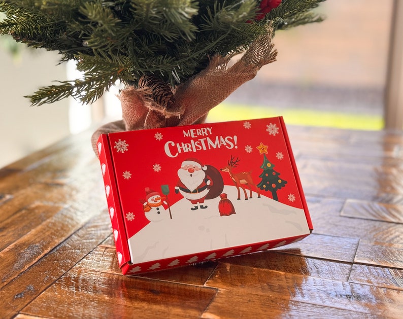 K&ouml;nnte beinhalten: Eine rechteckige rote Geschenkbox aus Karton mit dem Schriftzug Merry Christmas in wei&szlig;en Buchstaben. Die Box zeigt eine festliche grafische Illustration mit dem Weihnachtsmann, einem Schneemann, einem Rentier und einem Weihnachtsbaum.