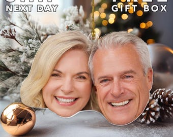 May include: A custom-shaped hanging ornament featuring a photographic portrait of a smiling blonde woman and a smiling man with gray hair. The ornament includes a gold hanging string and is displayed against a blurred holiday background.