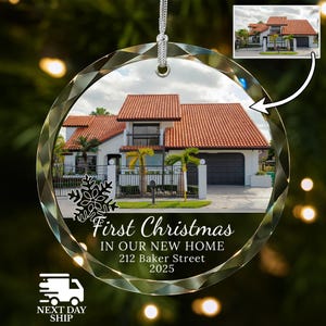 May include: A clear, faceted glass Christmas ornament with a silver string. The ornament features a house with a red tile roof and the text "First Christmas in Our New Home, 212 Baker Street, 2025."