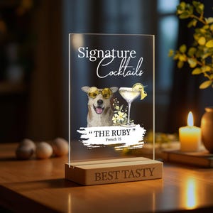May include: A clear acrylic sign with a cartoon dog wearing sunglasses and a cocktail glass. The sign reads "Signature Cocktails" and "The Ruby" with the text "French 75" underneath. The sign is on a wooden base with the text "Best Tasty" engraved on it.