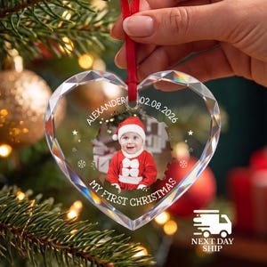 May include: A heart-shaped clear glass ornament with faceted edges, featuring a central photo of a baby in a red and white Santa outfit. The text reads ALEXANDER 02.08.2026 and MY FIRST CHRISTMAS, accented with small white stars and snowflakes.