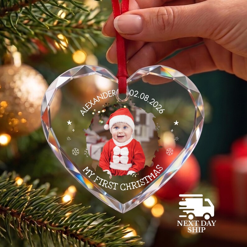 May include: A heart-shaped clear glass ornament with faceted edges, featuring a central photo of a baby in a red and white Santa outfit. The text reads ALEXANDER 02.08.2026 and MY FIRST CHRISTMAS, accented with small white stars and snowflakes.