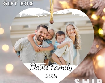 May include: A heart-shaped ceramic ornament featuring a printed photograph of a family of four. The text "Davis Family" is written in a black script font above the year "2024" in a simple serif font. It hangs from a gold cord.