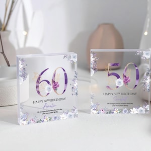 May include: Two clear acrylic blocks with floral designs and the numbers 60 and 50 in purple. The blocks read "Happy 60th Birthday Amelia" and "Happy 50th Birthday [Name]" with additional text. The floral accents include purple and white flowers.