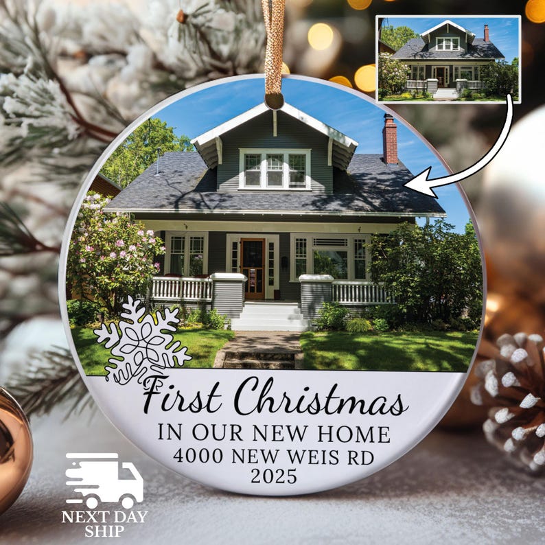 May include: A round Christmas ornament featuring a house photo, a snowflake, and the text "First Christmas In Our New Home 4000 New Weis Rd 2025." The ornament has a gold ribbon for hanging. The house is gray with white trim.