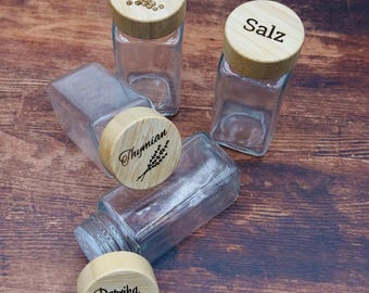 May include: Four clear glass spice jars with light-coloured wooden lids. The lids are engraved with the words Pfeffer, Salz, Thymian, and Paprika edelsüß, accompanied by small illustrations of peppercorns, a sprig of thyme, and a bell pepper.