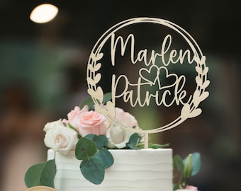 May include: A white wedding cake with a wooden cake topper that says "Marlen Patrick" with two hearts. The cake topper is surrounded by a wreath of leaves. A brown paper circle tag with the text "handmade with love" and a German flag is attached to the bottom tier of the cake.