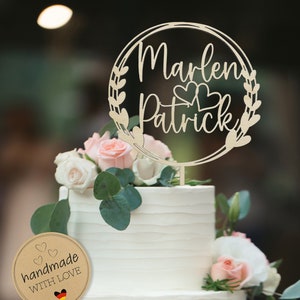 Könnte beinhalten: Eine weiße Hochzeitstorte mit einem Holz-Tortenaufsatz, auf dem "Marlen Patrick" mit zwei Herzen steht. Der Tortenaufsatz ist von einem Kranz aus Blättern umgeben. Ein braunes Papier-Rundetikett mit dem Text "handgefertigt mit Liebe" und einer deutschen Flagge ist an der untersten Schicht der Torte befestigt.