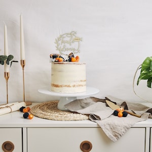 May include: A celebration cake with a gold-coloured topper, fresh fruit, and a white cake stand. The cake is on a white cabinet with candles, a plant in a woven basket, and a linen cloth. The cake is decorated with fresh fruit.