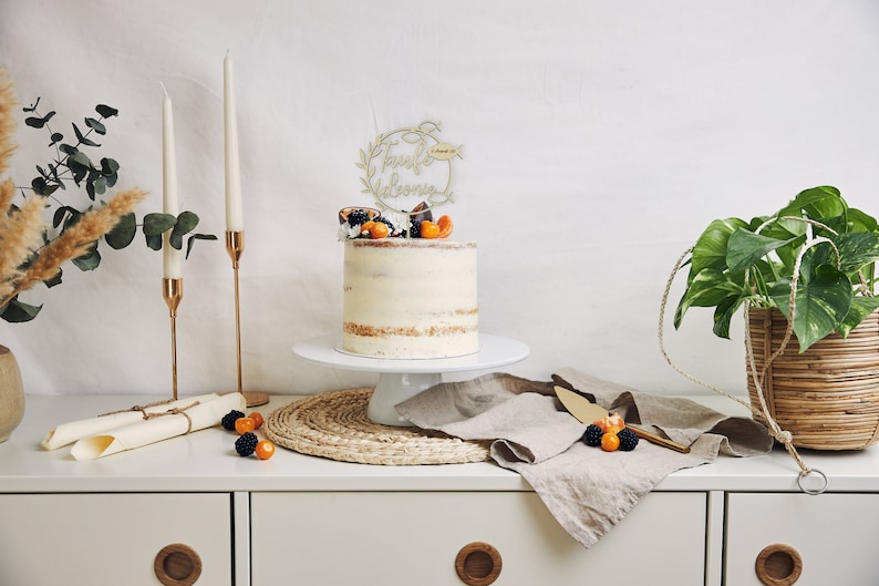 May include: A celebration cake with a gold-coloured topper, fresh fruit, and a white cake stand. The cake is on a white cabinet with candles, a plant in a woven basket, and a linen cloth. The cake is decorated with fresh fruit.